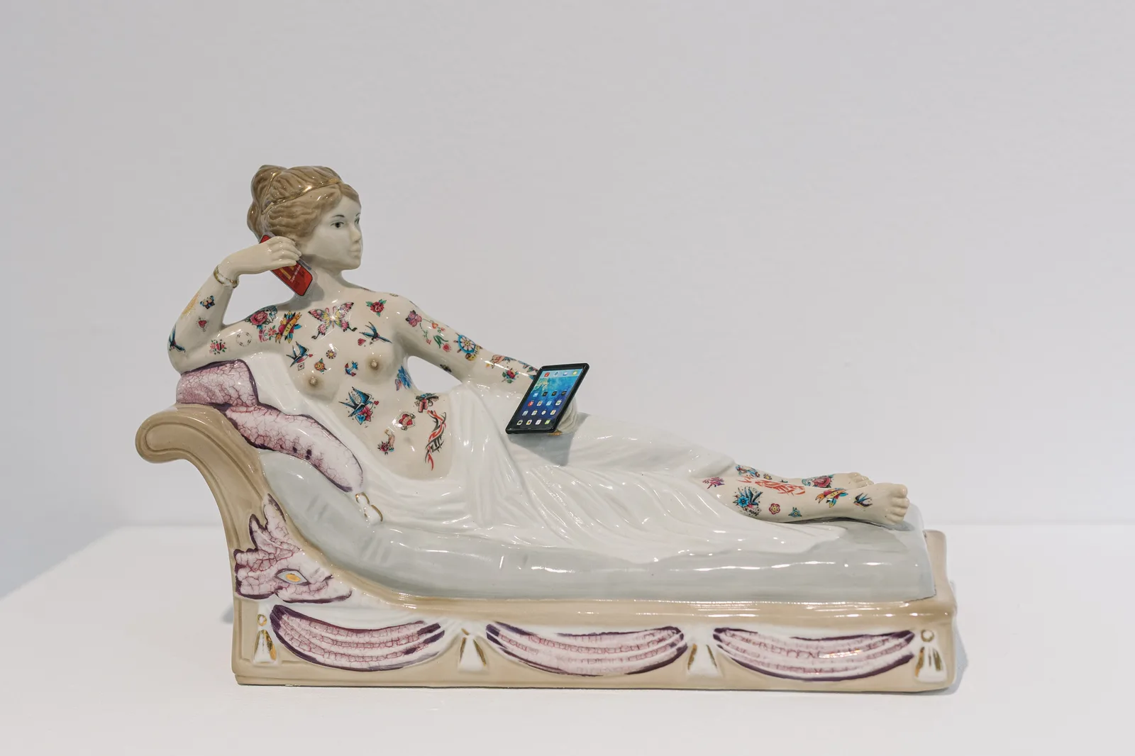 Glazed ceramic sculpture of a reclining classical female nude covered in colorful tattoos, holding a smartphone to her ear with a tablet propped on her hip, displayed on a white plinth against a white wall.