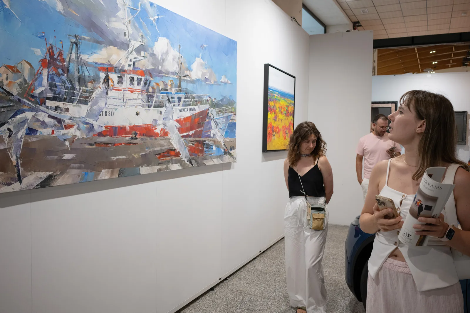 On the right stands a Tesla, and on the left hangs a painting. Two people in the background are looking at the Tesla, while the woman in the foreground is looking attentively at the painting.